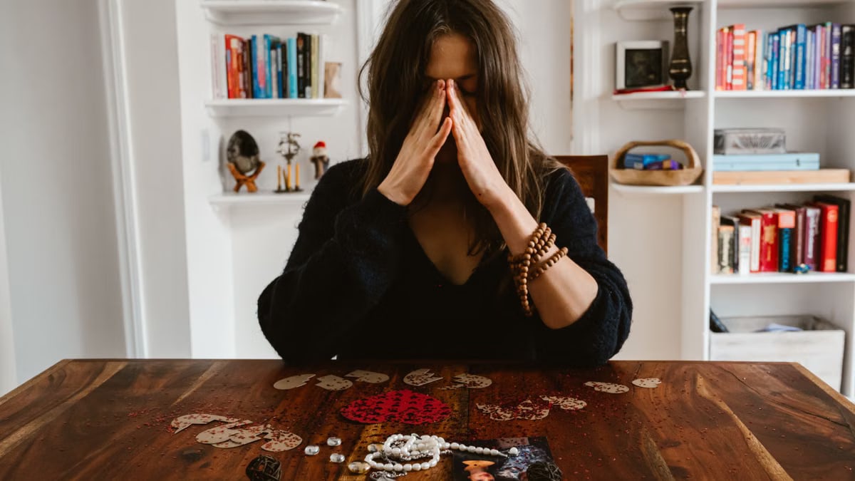 A woman in deep thought sits at a table with beads and a photo, conveying introspection.